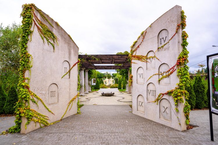 Dwei betonowe konstrukcje z wyrytymi ornammentami z numerami rzymskimi. Z tyłu drewniana pergola